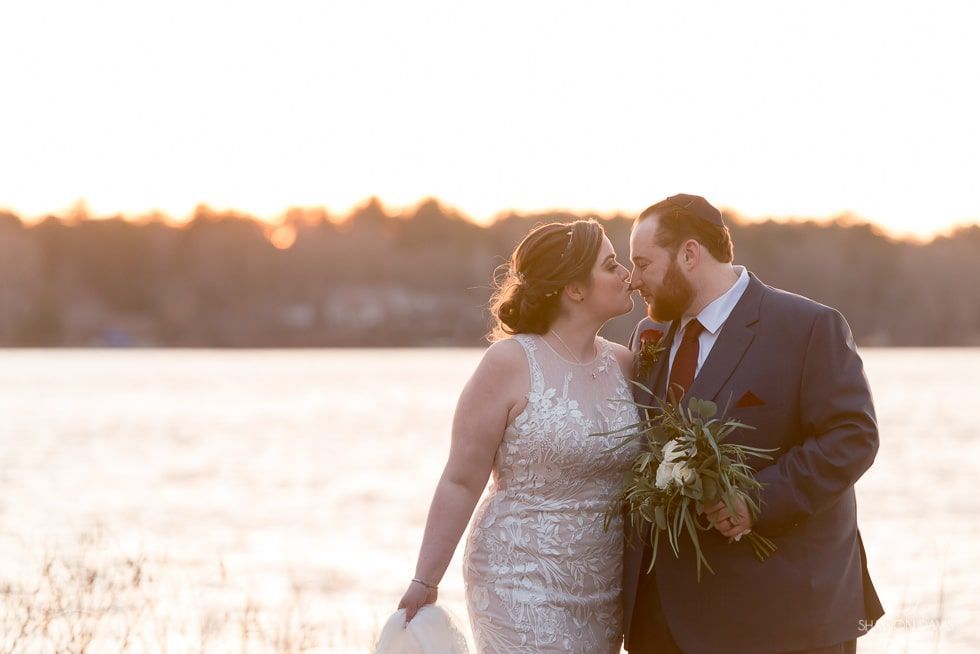 Couple in wedding attire kissing by a lake at sunset, with a bright sky and distant shoreline.
