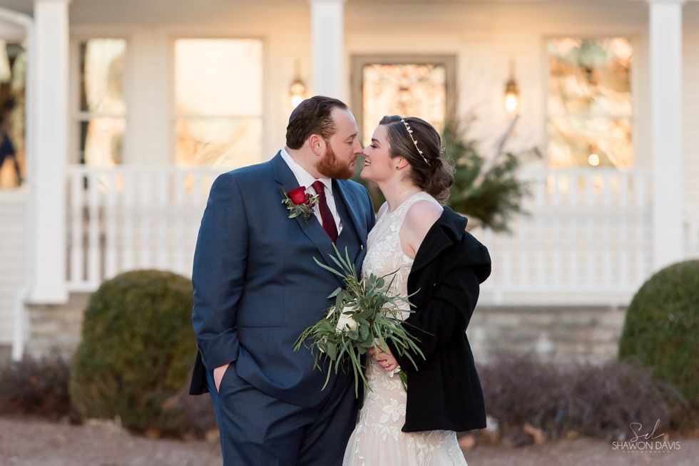 Two people in wedding attire share a kiss outdoors in front of a white house.