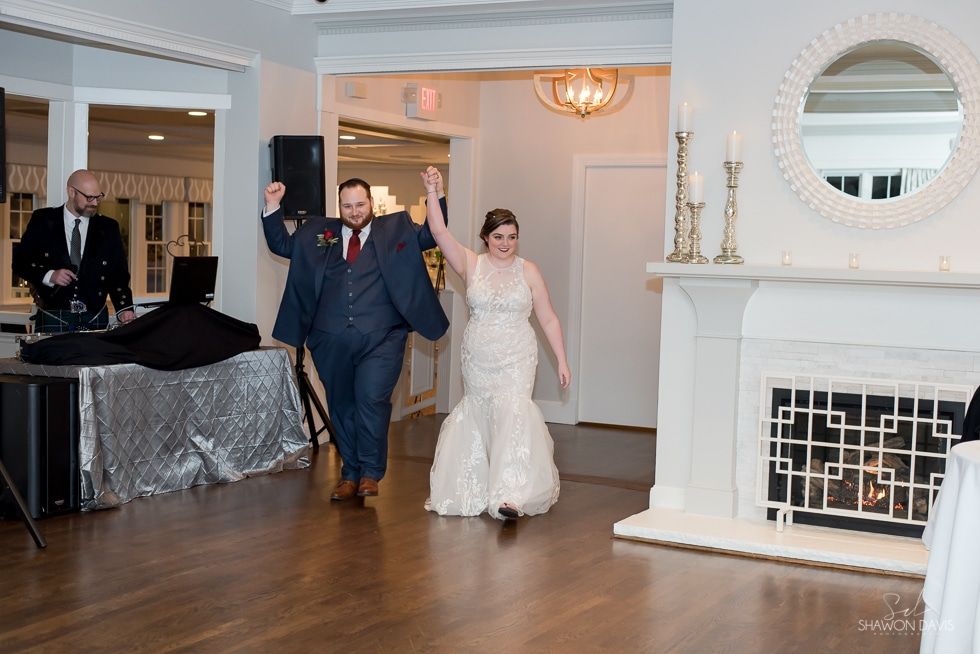 Bride and groom entering a ballroom, smiling and waving by a fireplace and DJ table