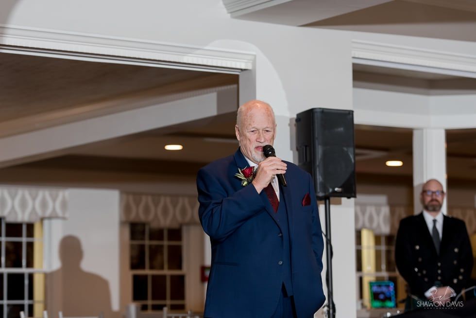 Man in a blue suit speaking into a microphone at an indoor event, with another man standing nearby