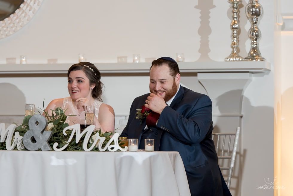 Bride and groom seated at a wedding reception table, smiling with drinks and “Mr. & Mrs.” decor