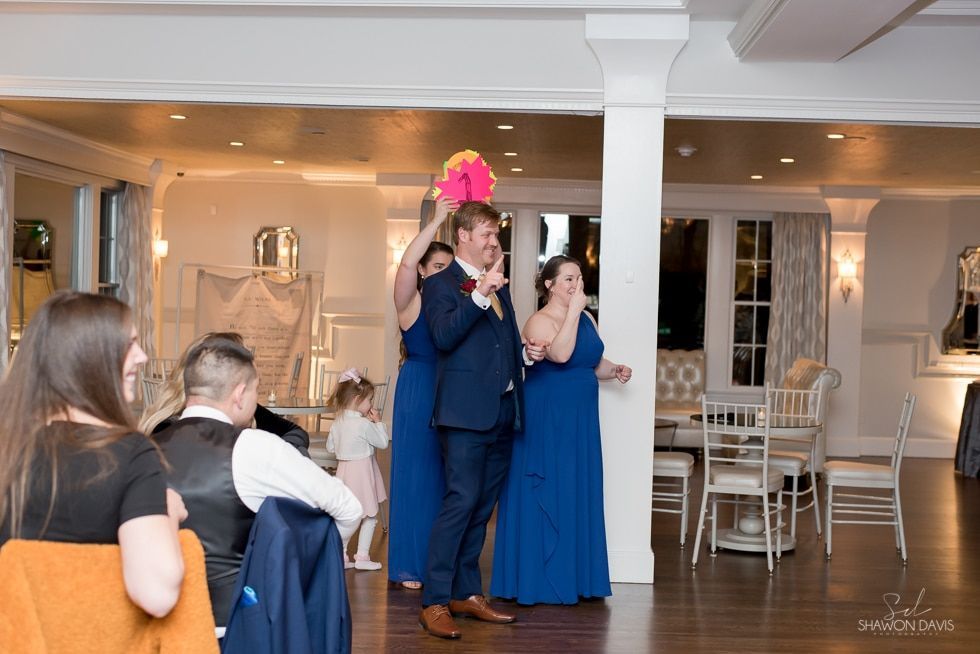 Two people in blue formal wear pose indoors at a reception, one holding up a bright pink prop.
