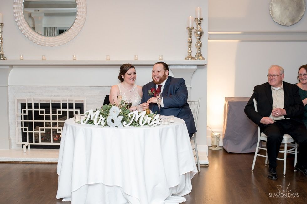 Wedding couple seated at a white table, smiling in a bright reception hall