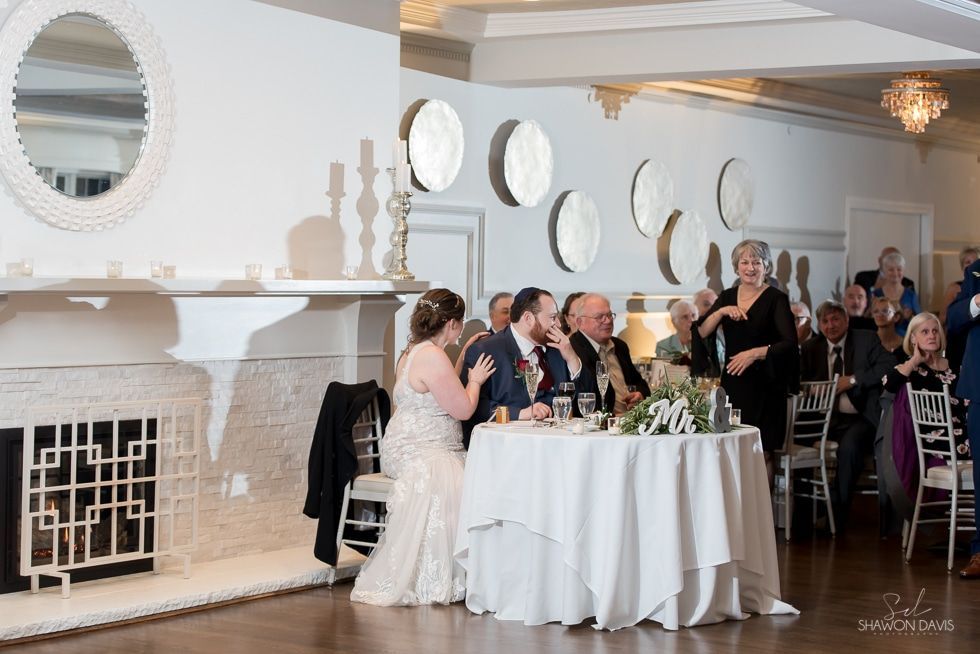 Wedding reception with bride, groom, and guests seated at a white-clothed table in a elegant hall