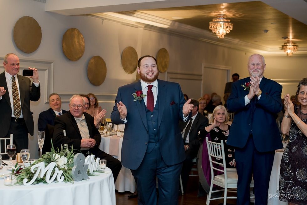 Man in navy suit smiling and dancing at a formal reception, with guests applauding nearby
