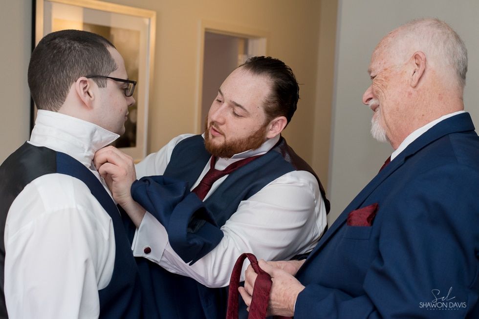 Two men adjusting a blue vest on a man in a white shirt indoors.