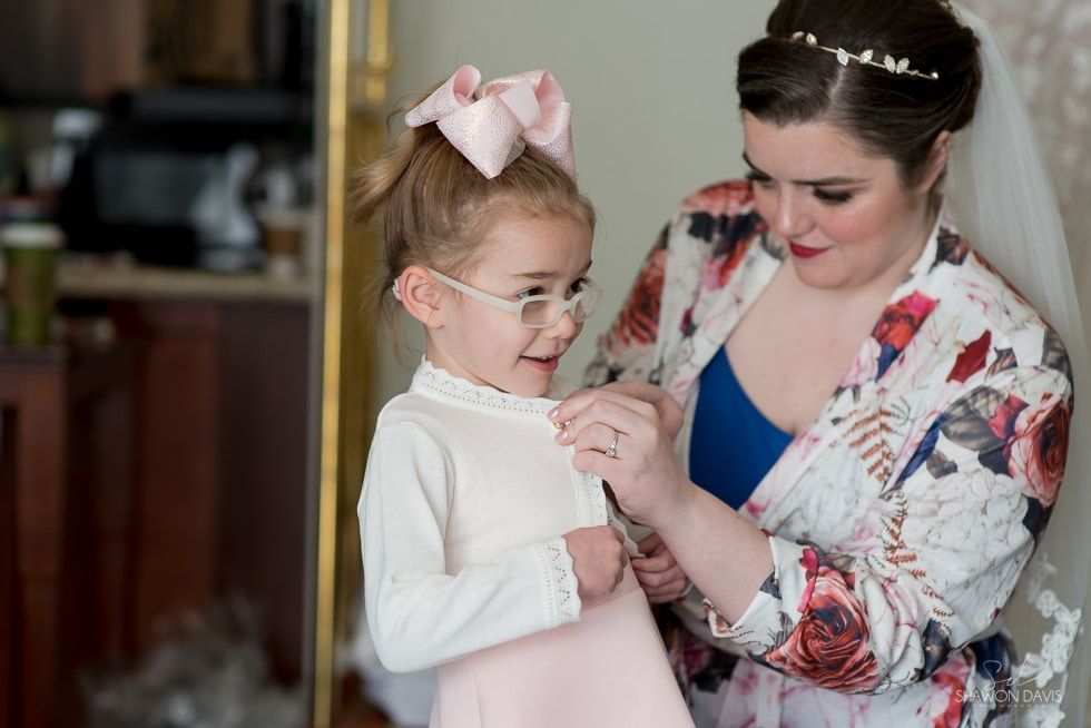 Woman adjusting a young girl’s dress and necklace, both dressed up indoors.