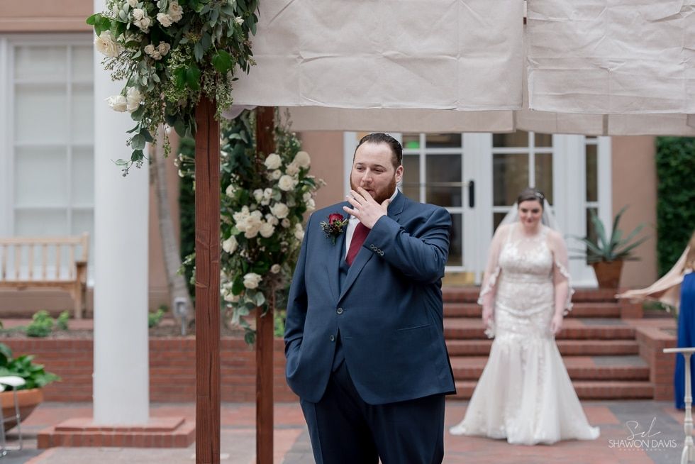 Man in navy suit wiping tears while bride in white dress stands behind him in a courtyard ceremony