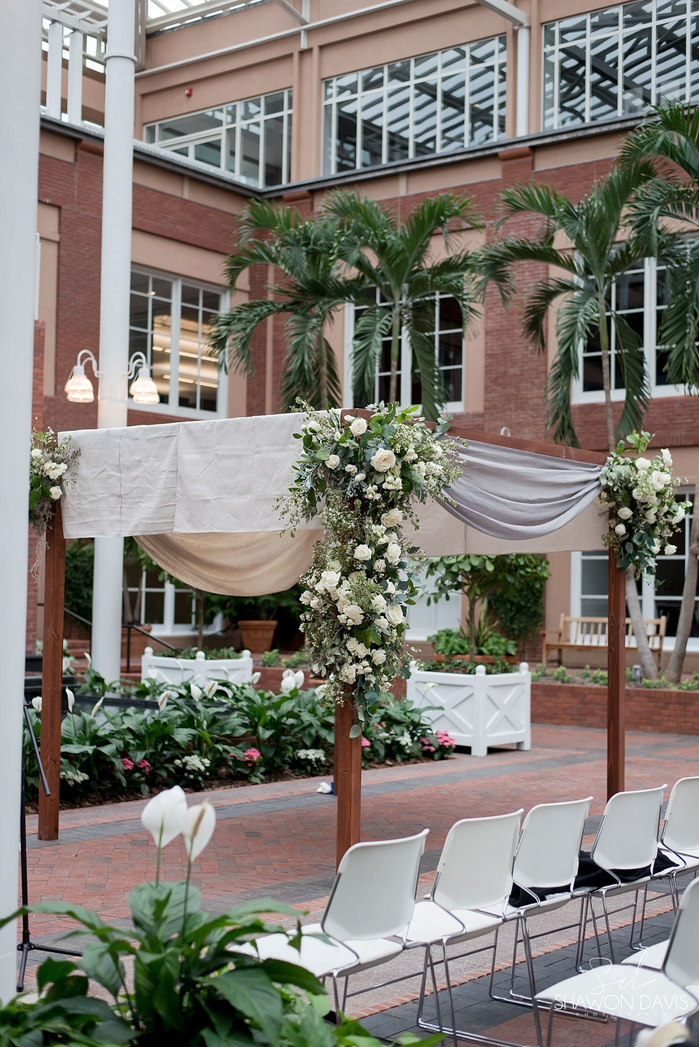 Decorated outdoor wedding setup with draped fabric, floral arrangements, and rows of white chairs in a brick courtyard