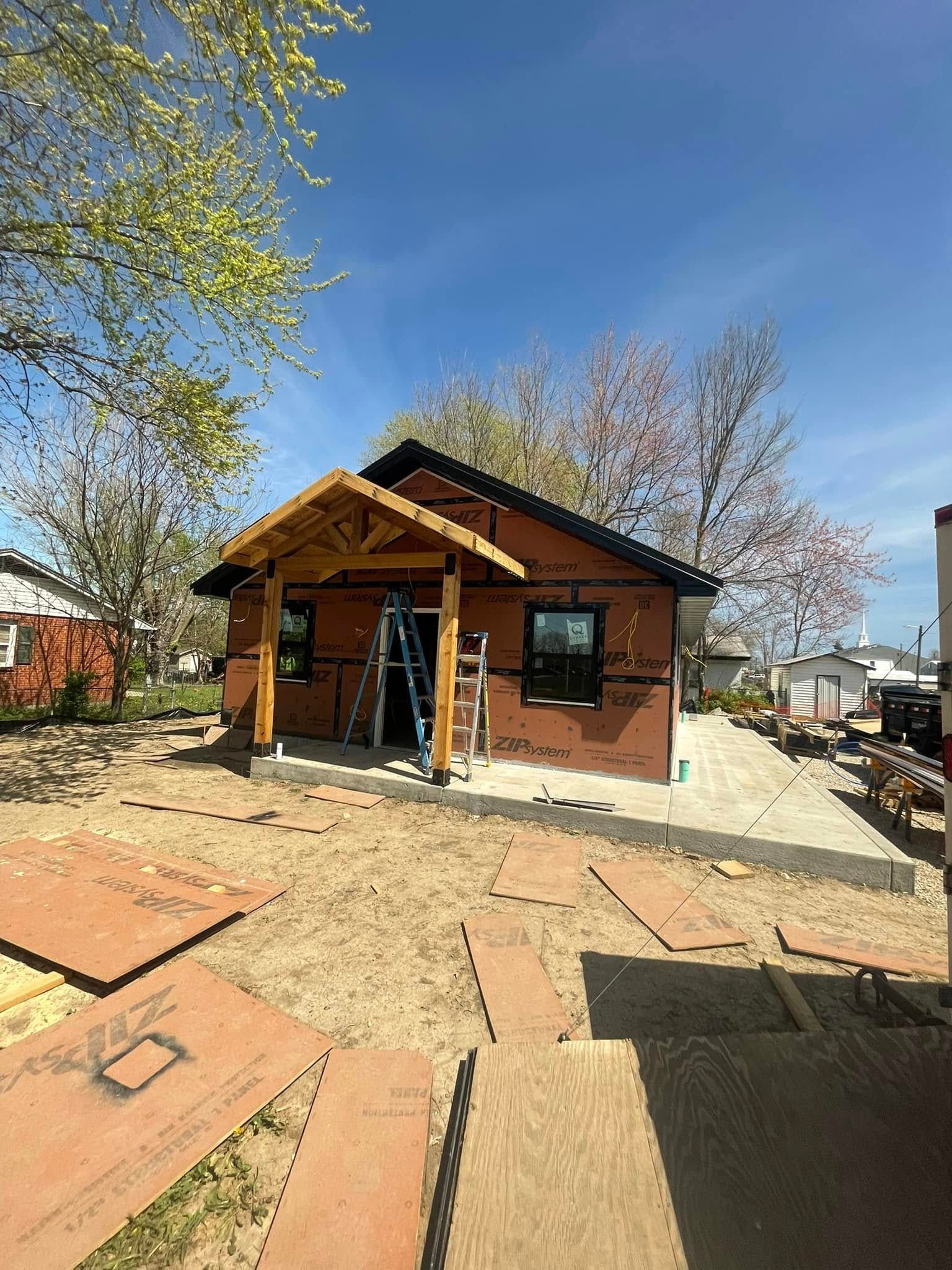 House under construction with a front porch, brown siding, and black roof, set on a sandy lot.
