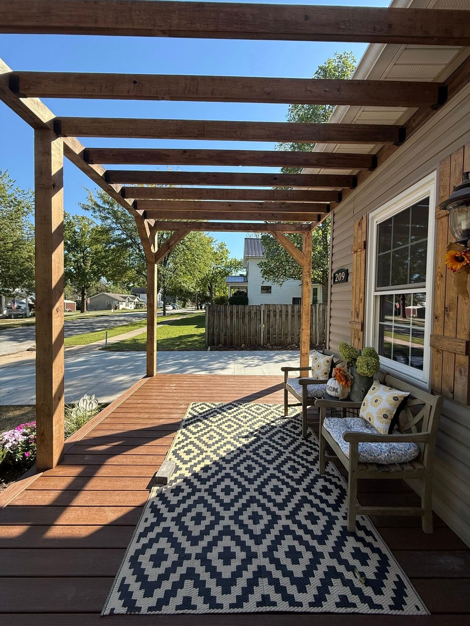 Wooden pergola over a porch with a patterned rug, chairs, and house siding.
