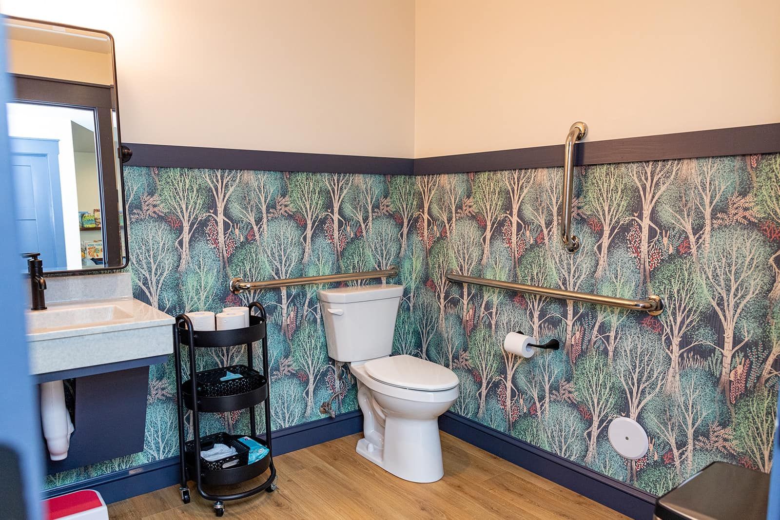 Accessible bathroom with forest wallpaper, toilet, sink, and grab bars.