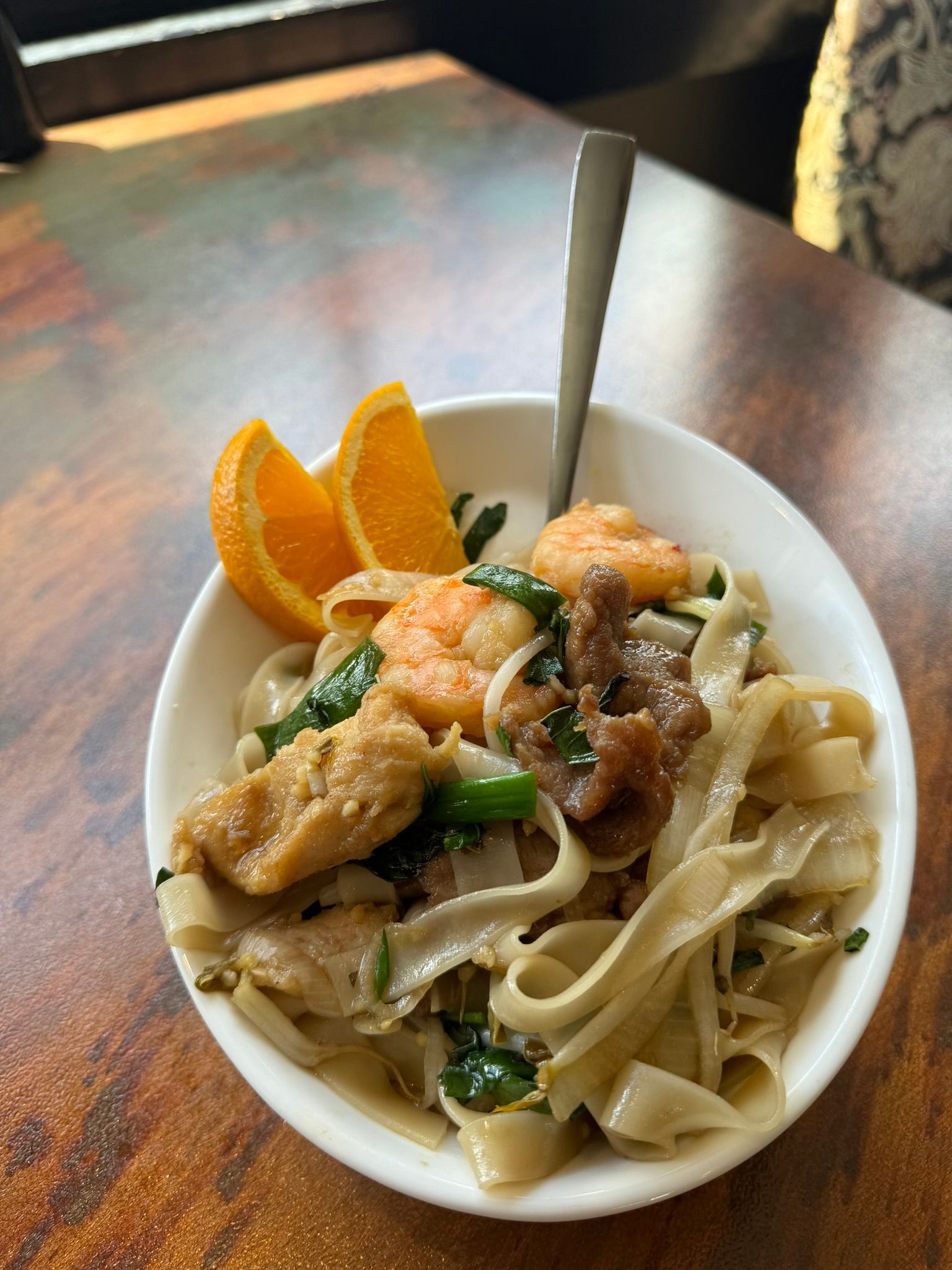 A bowl of food with noodles , meat , shrimp and oranges on a wooden table.