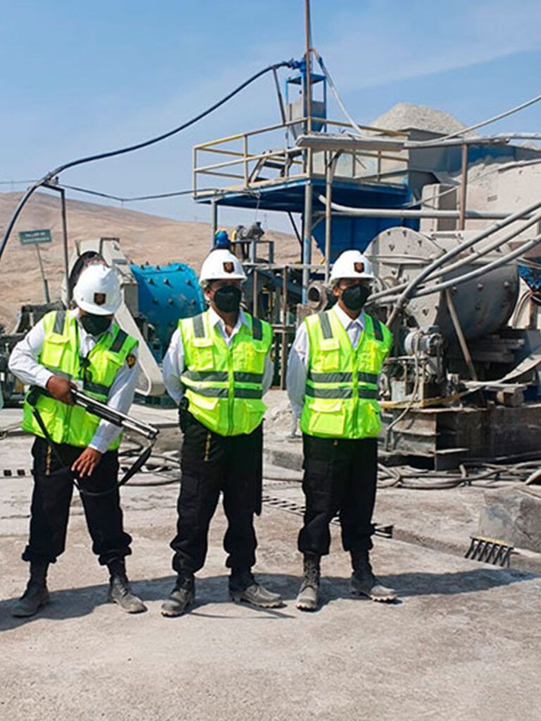 Tres guardias de seguridad con chalecos y máscaras se encuentran cerca de maquinaria en un sitio industrial al aire libre.