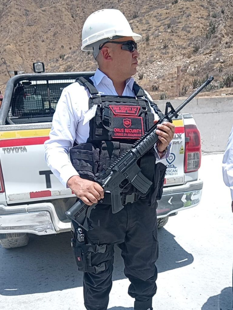 Un hombre con casco y equipo táctico sostiene un rifle cerca de una camioneta blanca en un entorno desértico.