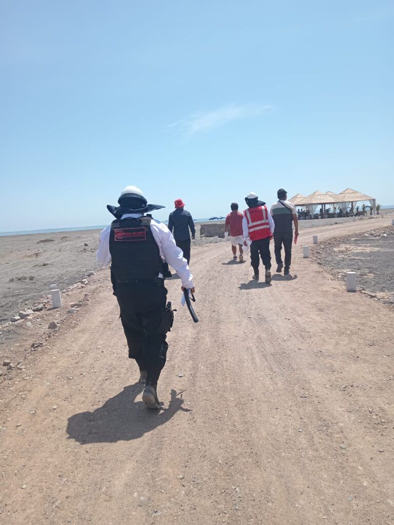 Una persona con equipo táctico camina por un sendero de tierra, seguida de otras; playa, cielo azul.