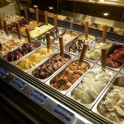 Vetrina espositiva di gelati con numerosi gusti in vaschette di metallo e palette di legno, in un negozio ben illuminato.