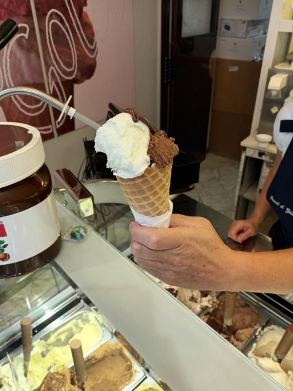 Una mano che regge un cono gelato con due palline di cialda al bancone di una gelateria.