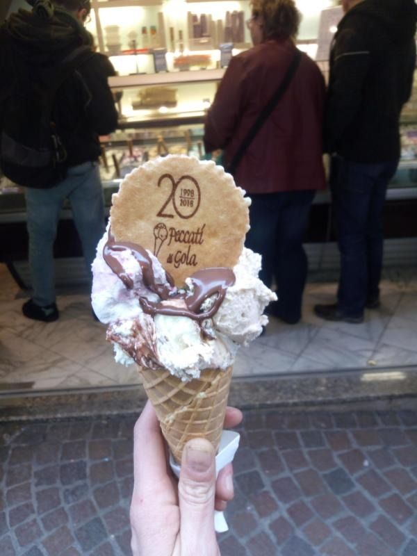Una mano regge un grande cono gelato ricoperto di panna montata, davanti al bancone di un negozio.