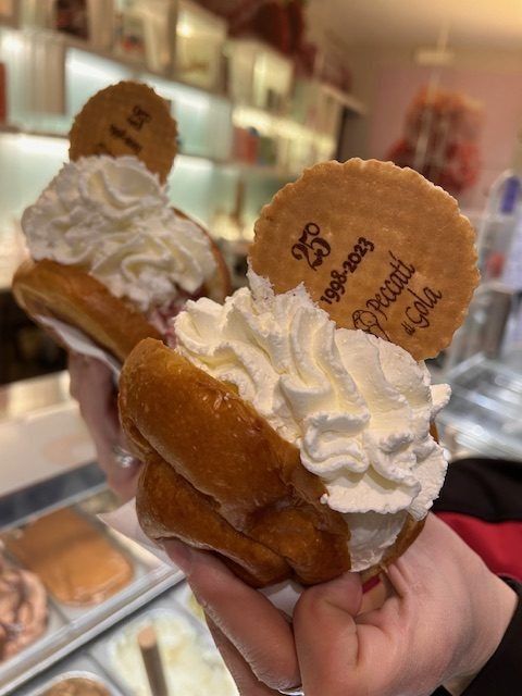 Due sandwich di gelato con panna montata e biscotti, tenuti in mano all'interno di una pasticceria.
