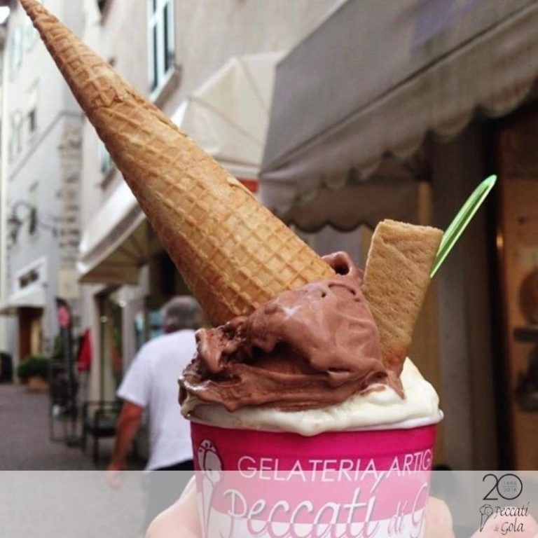 Gelato al cioccolato in una coppetta rosa con cono di cialda, ambientato in una strada cittadina.