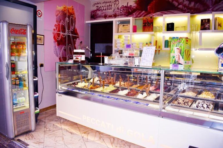 Bancone di una gelateria con vaschette colorate, vetrina e un frigorifero della Coca-Cola in un angolo.