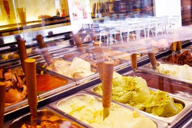 Vetrina per gelati con vari gusti e palette di legno in un negozio luminoso.