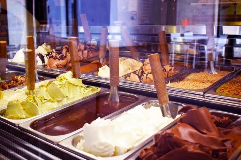 Assortimento di gusti di gelato in vassoi di acciaio inossidabile presso un bancone gelateria ben illuminato.