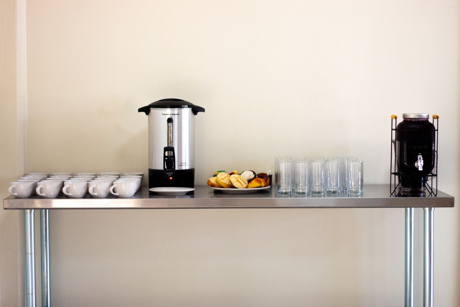 Una mesa de acero inoxidable con una cafetera, tazas y vasos sobre ella.