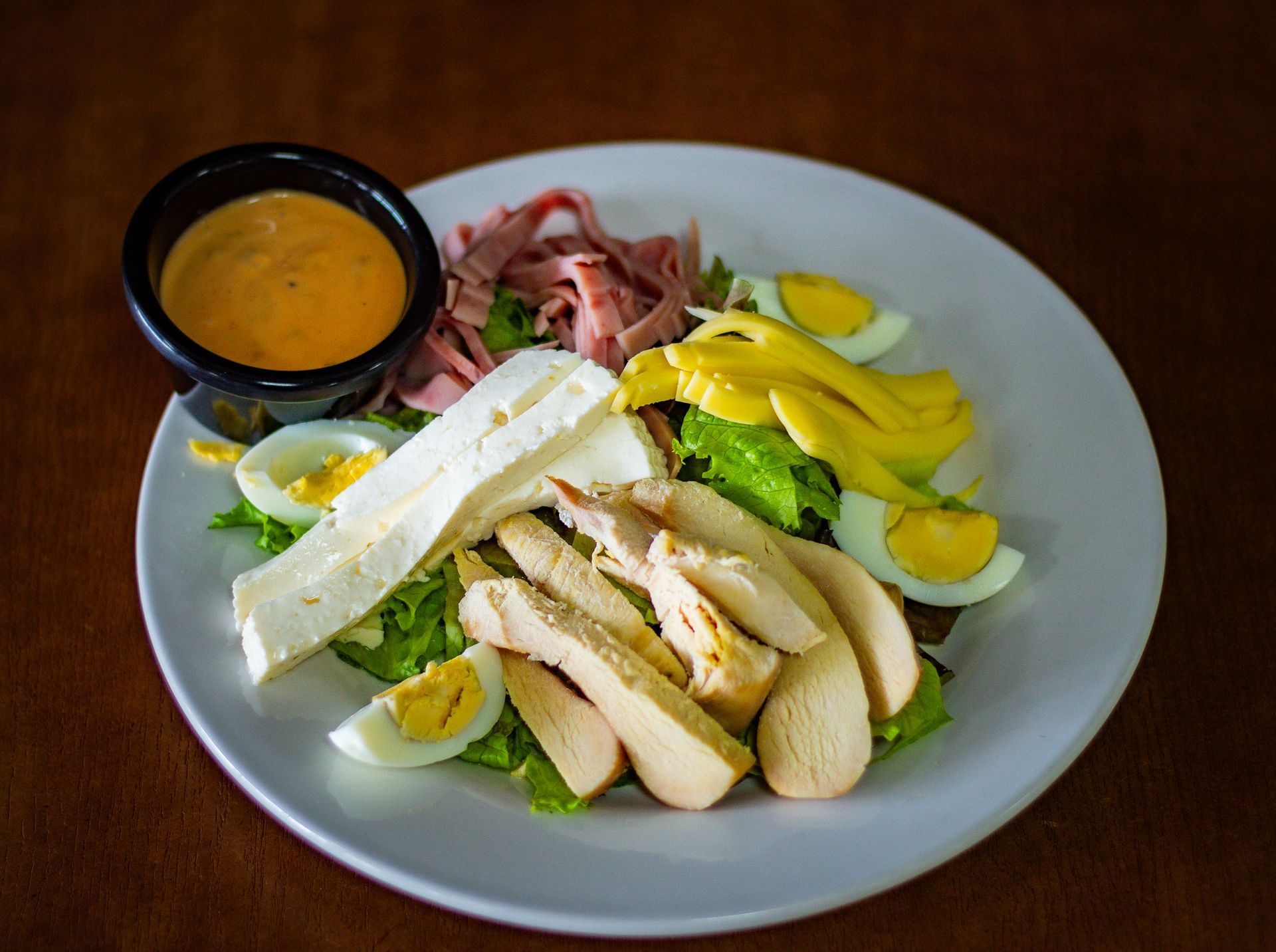 Una ensalada con pollo, huevos, jamón y queso en un plato con salsa para mojar.