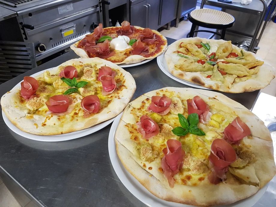 Quattro pizze su vassoi di metallo in una cucina, condite con pomodoro, formaggio, basilico e fette di salame piccante.