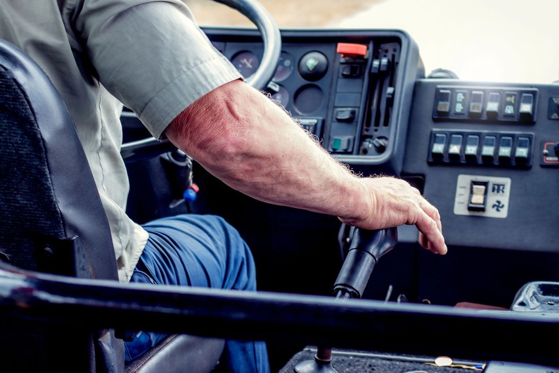 La mano di un autista è appoggiata sulla leva del cambio di un autobus, con i comandi del cruscotto visibili sullo sfondo.