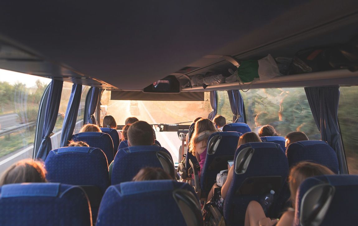 Vista dal retro di un autobus con passeggeri seduti su sedili blu, che guardano verso il parabrezza luminoso e illuminato dal sole.