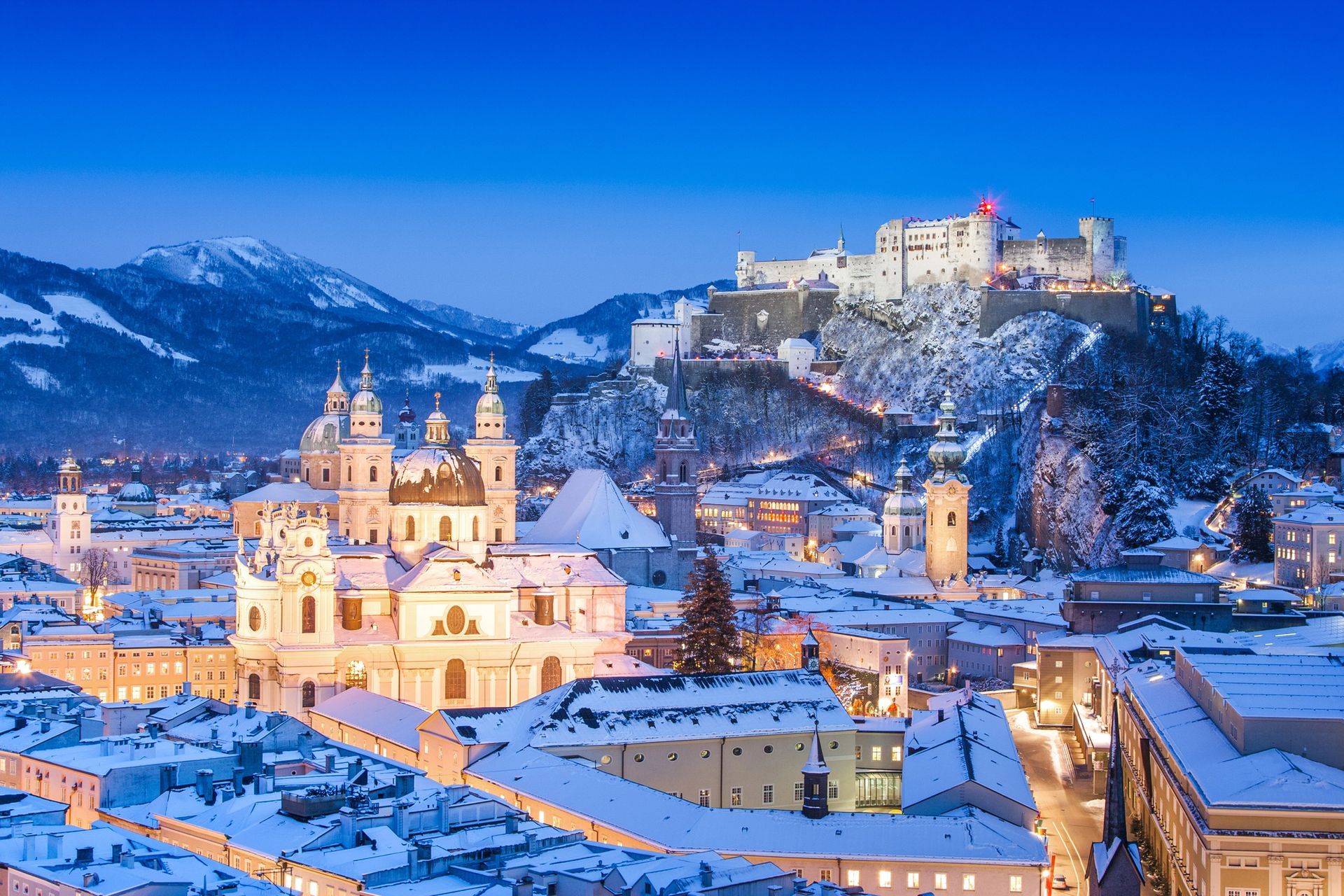 Salisburgo, Austria, al crepuscolo invernale, con la fortezza di Hohensalzburg illuminata che domina una città innevata.