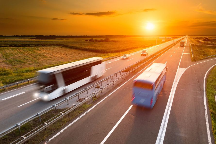 Un autobus bianco e un autobus blu percorrono un'autostrada a più corsie al tramonto, con la luce dorata che avvolge il paesaggio.