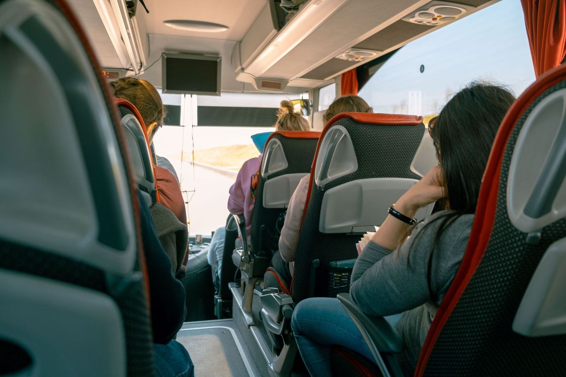 Passeggeri seduti in file all'interno di un autobus, visti da dietro, in viaggio verso un parabrezza luminoso e illuminato dal sole.