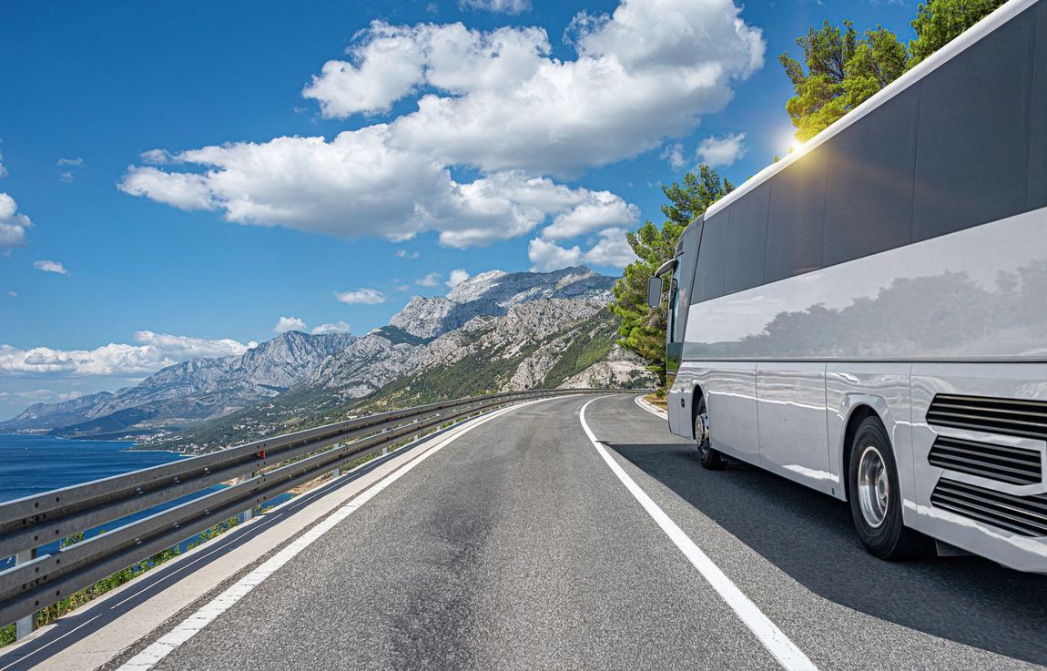 Un autobus turistico bianco percorre una strada costiera con le montagne sullo sfondo, sotto un cielo azzurro.