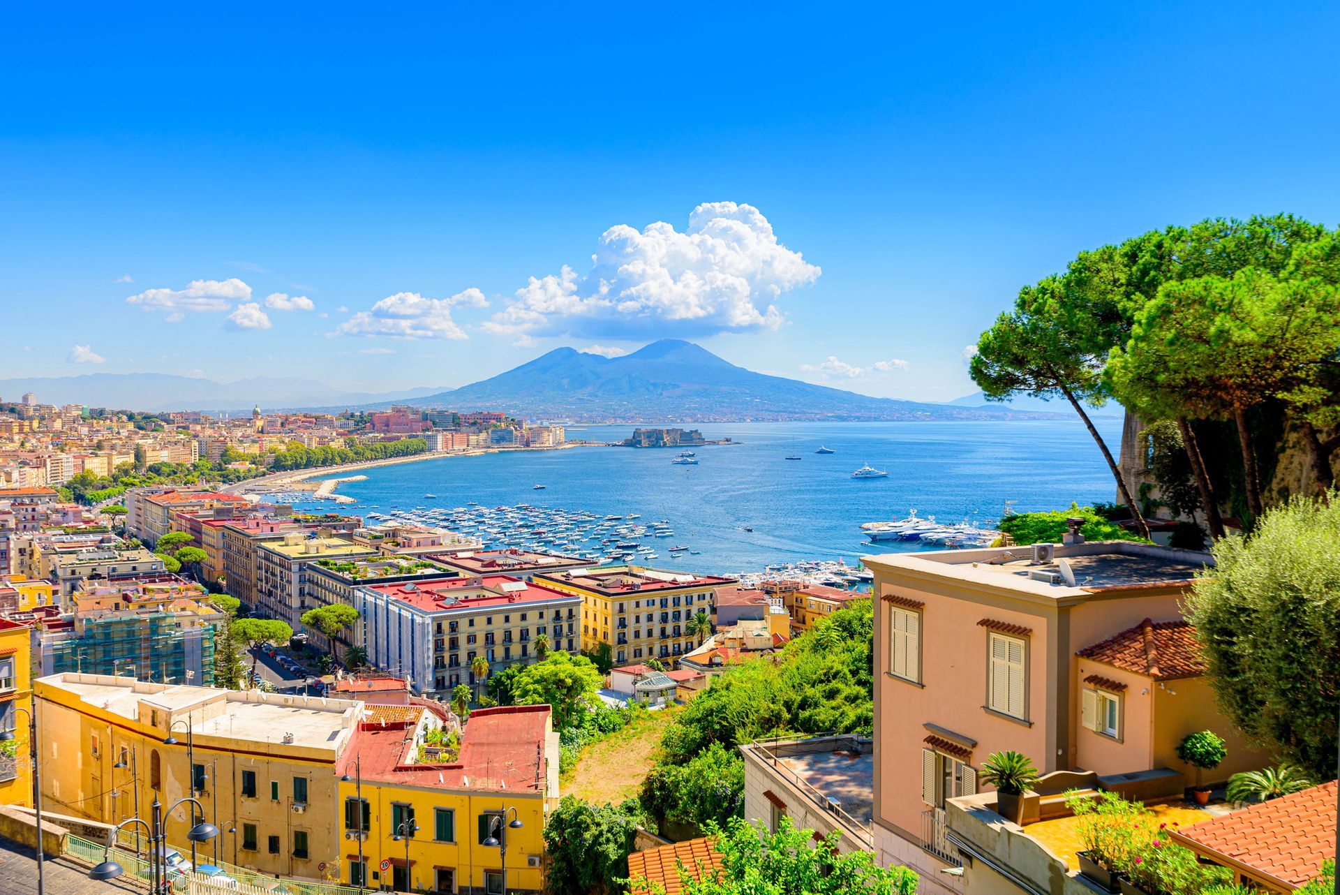Una vista soleggiata e dall'alto della costa della baia di Napoli, con il Vesuvio sullo sfondo, edifici colorati e alberi.