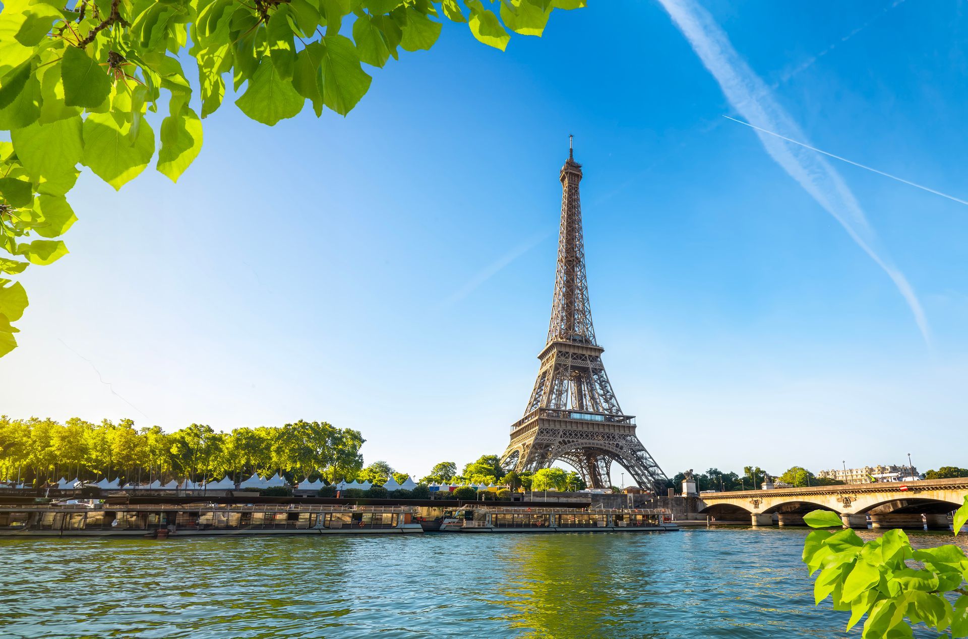 La Torre Eiffel si erge alle spalle di un fiume, sotto un cielo azzurro e limpido, incorniciata dal verde delle foglie in primo piano.