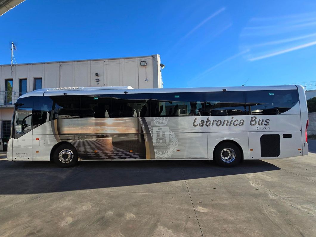 Un autobus Labronica bianco parcheggiato all'aperto, con una grande grafica raffigurante una piazza piastrellata su un fianco, sullo sfondo di un cielo azzurro limpido.