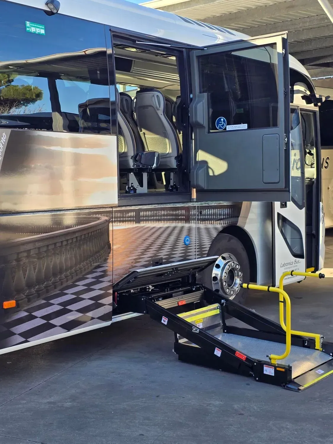 Un autobus con la porta laterale aperta e una piattaforma elevatrice per sedie a rotelle abbassata a livello del suolo.