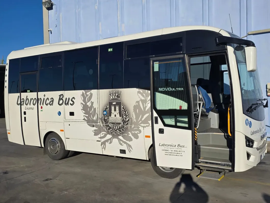 Un autobus bianco della Labronica parcheggiato all'aperto con la porta anteriore aperta, che mostra un emblema decorativo sul lato.