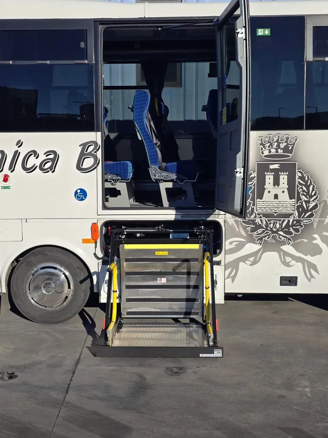Un autobus bianco con una porta laterale aperta e un elevatore nero per sedie a rotelle posizionato a livello del suolo per facilitare l'accesso.