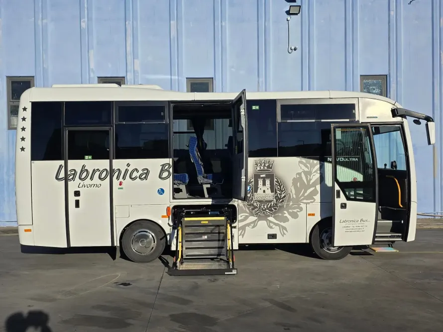 Un minibus bianco con la porta aperta e una piattaforma elevatrice per sedie a rotelle, parcheggiato di fronte a un edificio industriale blu.