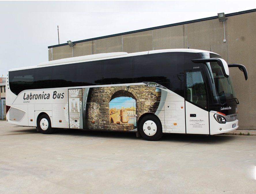 Un autobus Labronica bianco e nero parcheggiato su un piazzale asfaltato, con una grafica raffigurante rovine in pietra sul fianco.