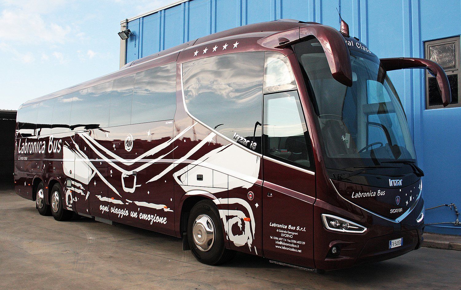 Un autobus Irizar i6 di colore rosso scuro con decorazioni grafiche bianche è parcheggiato all'aperto accanto a un edificio blu.