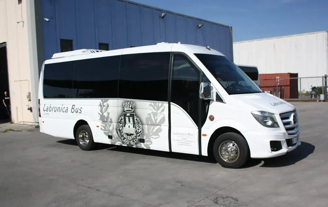 Un minibus Mercedes-Benz bianco con vetri oscurati parcheggiato su un piazzale asfaltato di fronte a un edificio industriale blu.