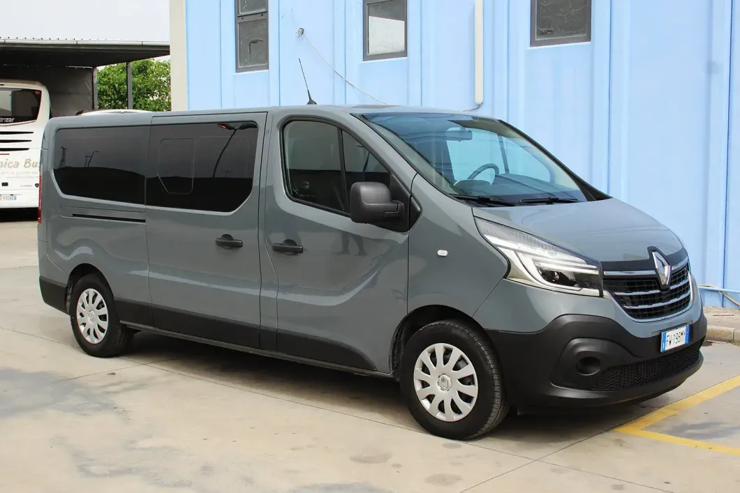 Un furgone passeggeri Renault Trafic grigio parcheggiato su un piazzale asfaltato di fronte a un edificio industriale azzurro.
