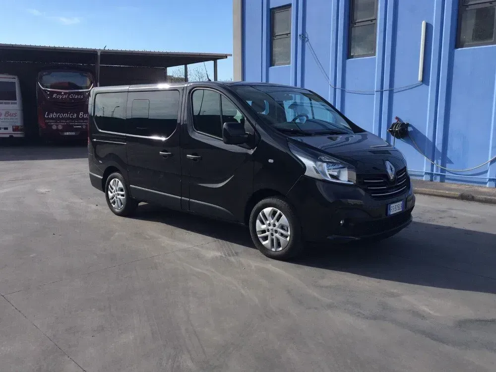 Un furgone passeggeri Renault Trafic nero parcheggiato su un piazzale asfaltato di fronte a un edificio blu, sotto un cielo sereno.