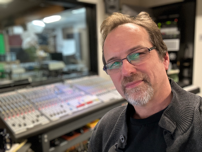 A man with glasses and a beard is standing in front of a sound board.